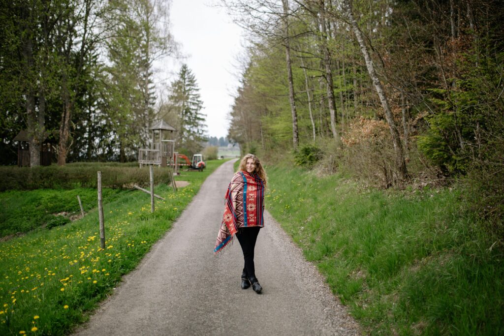 Woman with blonde hair in colorful shawl posing on a peaceful country road surrounded by spring greenery.